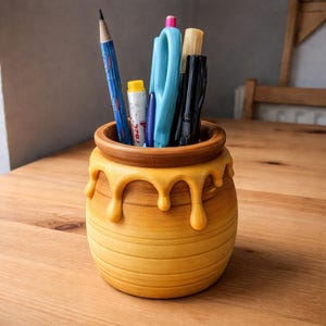 May include: A novelty desk tidy in the shape of a honey pot, filled with stationery. The pot is yellow with a dripping honey design. The pens and pencils are blue, black, and yellow. The organizer sits on a wooden table.