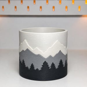May include: A cylindrical planter featuring a layered design with a white mountain peak top, a gray mid-section, and a black base decorated with a silhouette of pine trees. The surface has a fine, horizontal textured finish throughout the exterior.