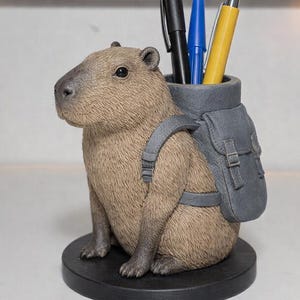 May include: A brown capybara figurine with a gray backpack, holding pens. The pen holder has a black base and contains a black pen, a blue pen, and a yellow pen. The capybara is sitting upright.