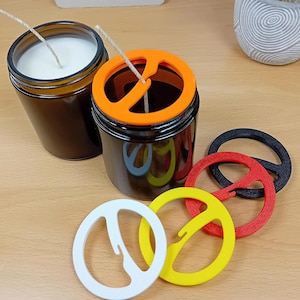 Può includere: Due barattoli di vetro marrone con cera di candela bianca e stoppini, uno con coperchio arancione. Quattro portacandele a forma di simbolo della pace colorati in bianco, giallo, rosso e nero sono accanto ai barattoli. I barattoli e i supporti sono su una superficie di legno marrone chiaro.