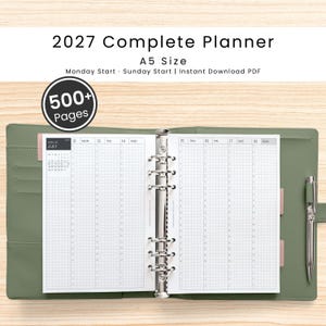 Op de afbeelding: Een open A5-planner voor 2027 met een saliegroene kaft en een zilverkleurig metalen ringbandmechanisme. De pagina's bevatten een weekindeling met een schema van maandag tot zondag, een kleine kalender voor juli en een 500+ pagina's indicator.