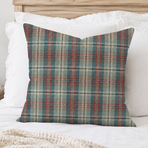 May include: A square throw pillow featuring a plaid pattern with intersecting lines in shades of muted teal, soft beige, and thin red stripes. The fabric has a woven texture, and the design consists of a repeating grid of vertical and horizontal lines.