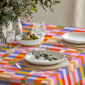May include: A rectangular tablecloth featuring a vibrant, multi-colored grid pattern of orange, pink, blue, yellow, and green squares. The table is set with white ceramic plates, clear glass stemware, and a bowl containing several green pears.