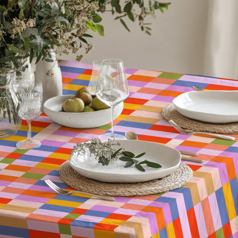 May include: A rectangular tablecloth featuring a vibrant, multi-colored grid pattern of orange, pink, blue, yellow, and green squares. The table is set with white ceramic plates, clear glass stemware, and a bowl containing several green pears.
