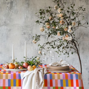 May include: A rectangular tablecloth featuring a vibrant, multi-colored checkered pattern in shades of orange, pink, blue, green, and yellow. The table is set with a white platter of assorted fresh apples, two white taper candles, and a stack of plates.