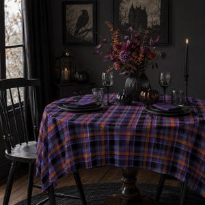 May include: A round table covered with a purple, orange, and black plaid tablecloth. The table is set with black plates, purple napkins, clear glasses, and a central vase filled with dark purple and orange flowers and foliage.