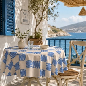 May include: A round table covered with a cream-colored tablecloth featuring a repeating pattern of blue fish. On the table sits a beige ceramic vase with green sprigs, a clear glass tumbler, and a small beige ceramic bowl.