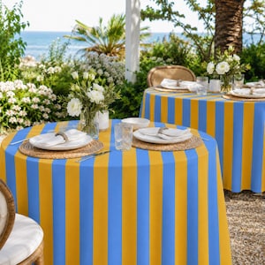 Può includere: Un tavolo rotondo coperto da una tovaglia a righe verticali blu e gialle. Il tavolo è apparecchiato con una tovaglietta intrecciata, un piatto bianco, un bicchiere trasparente e un vaso di vetro con fiori bianchi. È visibile un secondo tavolo.