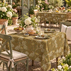 May include: A square olive green tablecloth featuring a white and yellow botanical pattern of leaves and small birds. The table is set with a ceramic pitcher of white hydrangeas, a bowl of pears, a loaf of bread, and plates with napkins.