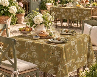 May include: A square olive green tablecloth featuring a white and yellow botanical pattern of leaves and small birds. The table is set with a ceramic pitcher of white hydrangeas, a bowl of pears, a loaf of bread, and plates with napkins.