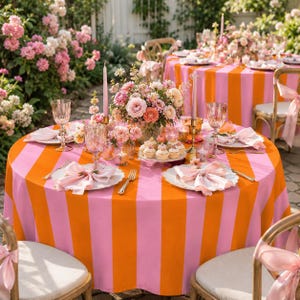 Op de afbeelding: Een ronde tafel bedekt met een roze en oranje gestreept tafelkleed, gedekt met witte borden, roze servetten, kristallen glazen, gouden bestek en een centraal bloemstuk met roze en perzikkleurige rozen en kleine cupcakes.