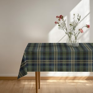 May include: A rectangular tablecloth featuring a dark blue, forest green, and beige plaid pattern. Two clear glass vases sit on the surface, each containing a small arrangement of pink carnations and white baby's breath flowers.