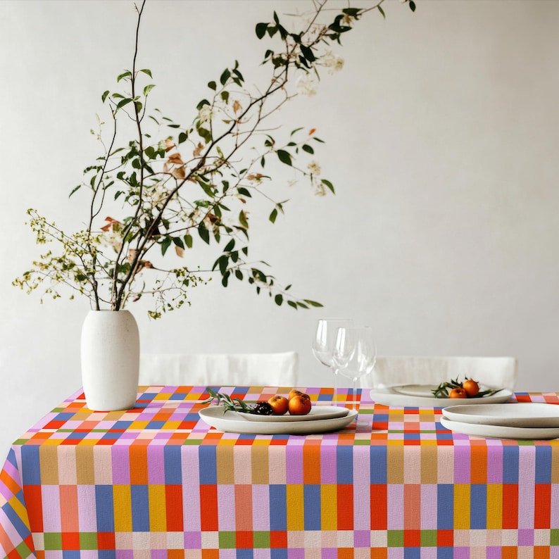 May include: A rectangular tablecloth featuring a vibrant grid pattern of orange, light blue, lavender, yellow, and green vertical and horizontal stripes. The tablecloth is set with white ceramic plates and clear glass stemware.