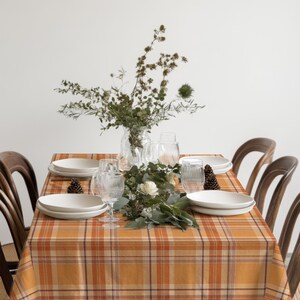 May include: A dining table covered in an orange, brown, and white plaid tablecloth, set with white ceramic plates, clear ribbed glassware, and a central arrangement of green foliage, dried branches, and pinecones, surrounded by wooden chairs.