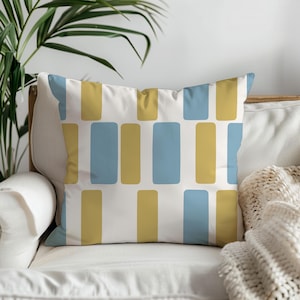 Op de afbeelding: Een vierkant decoratief kussen met een herhalend patroon van blauwe en gouden rechthoeken op een witte achtergrond. Het kussen ligt op een witte bank, met een plant op de achtergrond.