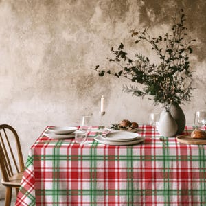 May include: A rectangular tablecloth featuring a classic plaid pattern in vibrant red, forest green, and crisp white stripes. The fabric covers a dining table set with white ceramic plates, clear glassware, a tall candle, and a vase with dried foliage.