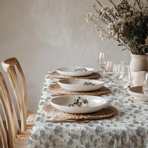 May include: A dining table set for a meal, featuring white ceramic plates on woven placemats. The table is covered with a floral patterned tablecloth. Clear wine glasses and a vase of dried flowers complete the setting. Wooden chairs are visible.