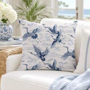 May include: A square throw pillow featuring a repeating pattern of blue cranes in flight against a light cream background. The design includes detailed illustrations of the birds with outstretched wings and stylized clouds, creating a cohesive visual.