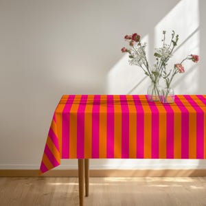 Puede incluir: Un mantel rectangular con rayas verticales en rosa brillante y naranja. El mantel está sobre una mesa de madera. Dos jarrones de cristal con flores están sobre la mesa.