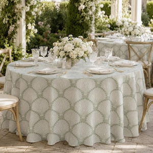 May include: A round table covered with a light green tablecloth featuring a repeating white fan-shaped shell pattern. The table is set with clear glass stemware, white ceramic plates, folded white napkins, and a central bouquet of white flowers.