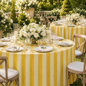 May include: A round table covered with a yellow and white striped tablecloth, set with white plates, clear glassware, and a central floral arrangement of white roses and greenery, surrounded by wooden cross-back chairs with white cushions.