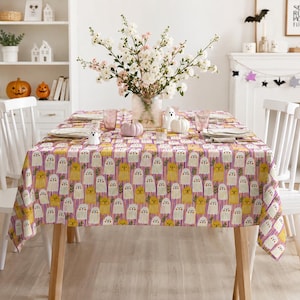 May include: A rectangular tablecloth featuring a repeating pattern of white and yellow ghosts on a background of vertical purple and white stripes. The tablecloth is draped over a wooden table set with white plates, pink glasses, and a glass vase of flowers.