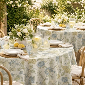 May include: A round tablecloth featuring a blue and green floral pattern on a white background, covering a table set with white plates, clear glassware, a glass pitcher of lemonade with lemon slices, and a white ceramic vase of flowers.