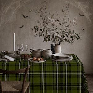 May include: A green, black, and white plaid tablecloth covers a table set with stacked ceramic bowls, plates, a tall white candle, two wine glasses, a small bowl of apples, and a vase containing bare branches with a black bird figurine.