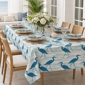 May include: A rectangular tablecloth featuring a repeating pattern of dark blue herons on a light blue and white textured background. The table is set with white plates, woven placemats, blue glassware, and a large bouquet of white flowers.