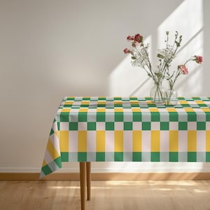 May include: A rectangular tablecloth featuring a repeating geometric pattern of alternating green, yellow and light pink squares and rectangles. Two clear glass vases containing small pink and white flowers sit on the surface of the table.