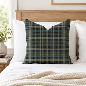 May include: A square throw pillow featuring a plaid pattern in shades of dark blue, forest green, and tan. The fabric displays a classic intersecting grid design with thin black lines running horizontally and vertically across the surface.