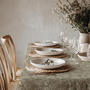 May include: A dining table set with a sage green patterned tablecloth, three white ceramic bowls placed on woven round placemats, clear glass stemware, and a white vase filled with dried white flowers, accompanied by wooden chairs.