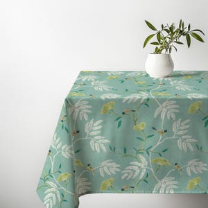Può includere: Una tovaglia rettangolare color verde acqua tenue con un motivo ripetuto di foglie bianche simili a felci, sottili rami marroni, piccoli fiori gialli e minuscoli uccellini con testa blu e petto giallo, sormontata da un vaso bianco.