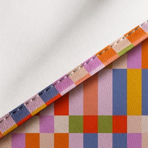 May include: A textile fabric featuring a repeating geometric pattern of vertical rectangular blocks in shades of orange, yellow, lavender, blue, and green. The fabric has a clean, white finished edge with visible white stitching along the seam.