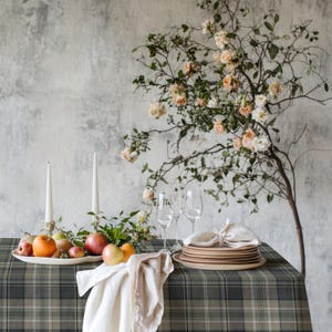 May include: A dining table set for a special occasion. The table is covered with a green and gray plaid tablecloth. It is set with plates, glasses, and a white linen napkin. A white platter of fruit and two white candles are on the table. A floral arrangement is in the background.