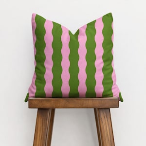 May include: A square decorative pillow with a wavy striped pattern in olive green and light pink. The pillow is placed on a wooden stool with a light background.