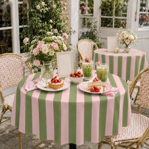 Peut inclure: Une table ronde recouverte d'une nappe à rayures roses et vert sauge, avec un vase en verre rempli de fleurs roses, un petit gâteau aux fraises, un bol de fraises et un verre de boisson glacée verte sur une assiette blanche.