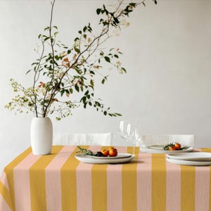 May include: A dining table set with a striped tablecloth in mustard yellow and pale pink. The table is set with white plates, wine glasses, and a white vase with branches and leaves. Two small oranges and berries are on a plate.