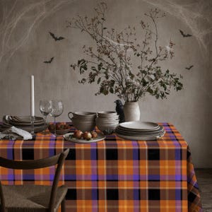 May include: A rectangular tablecloth featuring a vibrant plaid pattern in shades of orange, black, and purple. The fabric is draped over a table set with stacks of neutral-toned ceramic bowls, clear glass stemware, a white candle, and a vase.
