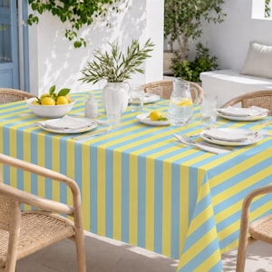 Könnte beinhalten: Eine rechteckige Tischdecke mit vertikalen gelben und hellblauen Streifen. Der Tisch ist mit weißen Tellern, gefalteten weißen Servietten, einer gläsernen Wasserkaraffe, einer weißen Vase mit Grünpflanzen und einer Schale mit gelben Zitronen gedeckt.