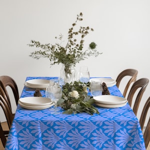 May include: A rectangular tablecloth featuring a repeating fan-shaped pattern in shades of bright blue and light periwinkle. The table is set with four white ceramic plates, four clear ribbed drinking glasses, and a central arrangement of greenery.
