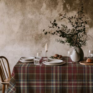 May include: A rectangular tablecloth featuring a dark red, brown, and blue plaid pattern. The table is set with white ceramic plates, clear glass stemware, a single white taper candle in a holder, and a vase containing dried foliage.