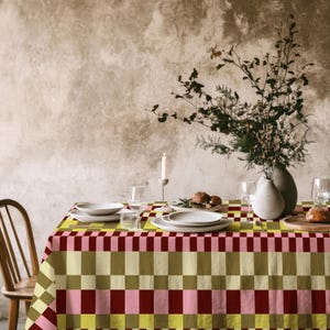 Può includere: Una tovaglia rettangolare caratterizzata da un motivo geometrico a scacchiera ripetuto in tonalità di verde oliva spento, rosso bordeaux intenso e rosa pastello tenue. Il tessuto copre un tavolo da pranzo apparecchiato con piatti in ceramica.