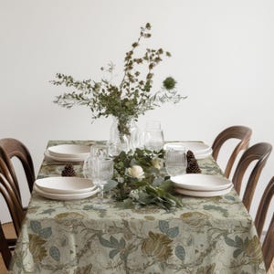 Puede incluir: Una mesa de comedor rectangular cubierta con un mantel verde claro que presenta un patrón floral detallado en tonos marrón, azul y verde. La mesa está dispuesta con platos de cerámica blanca, cristalería estriada transparente y un centro floral.