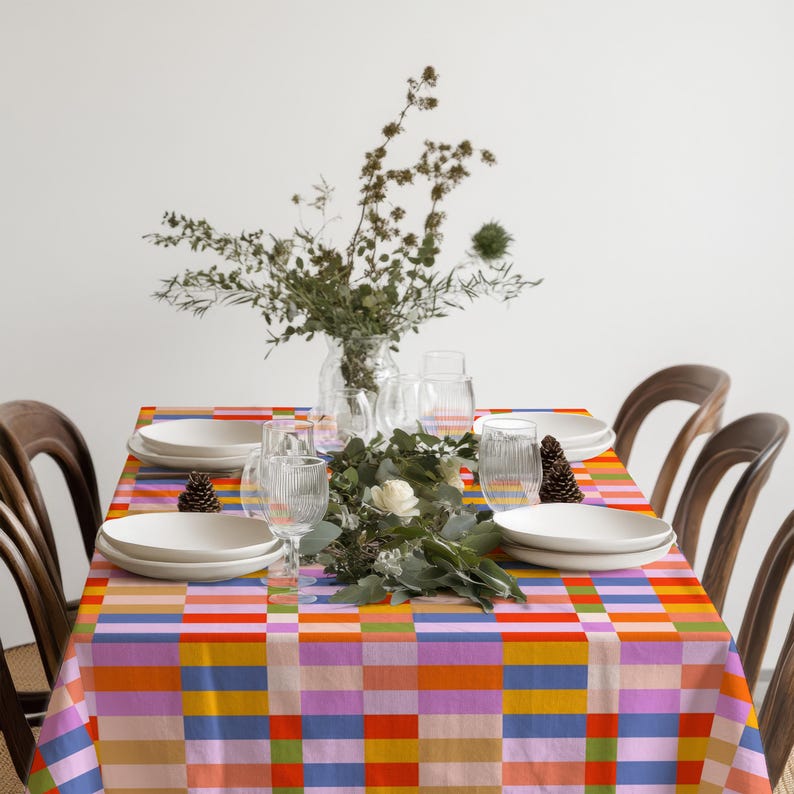 May include: A rectangular tablecloth featuring a vibrant grid pattern of alternating blocks in orange, blue, pink, yellow, and green. The fabric is draped over a dining table set with white plates, clear ribbed glasses, and a central floral arrangement.