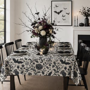 May include: A dining table set with a black and white floral tablecloth featuring bat silhouettes. The table includes black chairs, place settings with black napkins, clear glassware, and a central vase filled with dark flowers and bare branches.