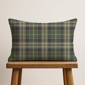 May include: Rectangular decorative pillow with a green, navy, and tan plaid pattern. The pillow is placed on a wooden bench. The plaid design features a classic tartan style, with intersecting lines creating a grid-like pattern.