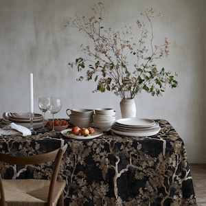 Puede incluir: Una mesa de comedor cubierta con un mantel negro con un patrón de árboles en color canela, con una pila de platos y cuencos de cerámica blanca, dos copas de vino transparentes, una vela blanca en un portavelas, un cuenco de bayas y un jarrón.