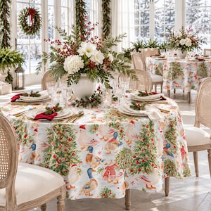 May include: A round tablecloth featuring a festive print of ducks wearing winter accessories and decorated Christmas trees. The table is set with white plates, clear glassware, gold cutlery, and a centrepiece of white flowers and red berries.