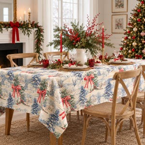 Puede incluir: Un mantel rectangular con una escena invernal festiva de árboles de hoja perenne azules, casas blancas y lazos rojos estampados sobre un fondo crema. La tela cubre una mesa de madera rodeada de sillas de madera clara con respaldo en cruz.